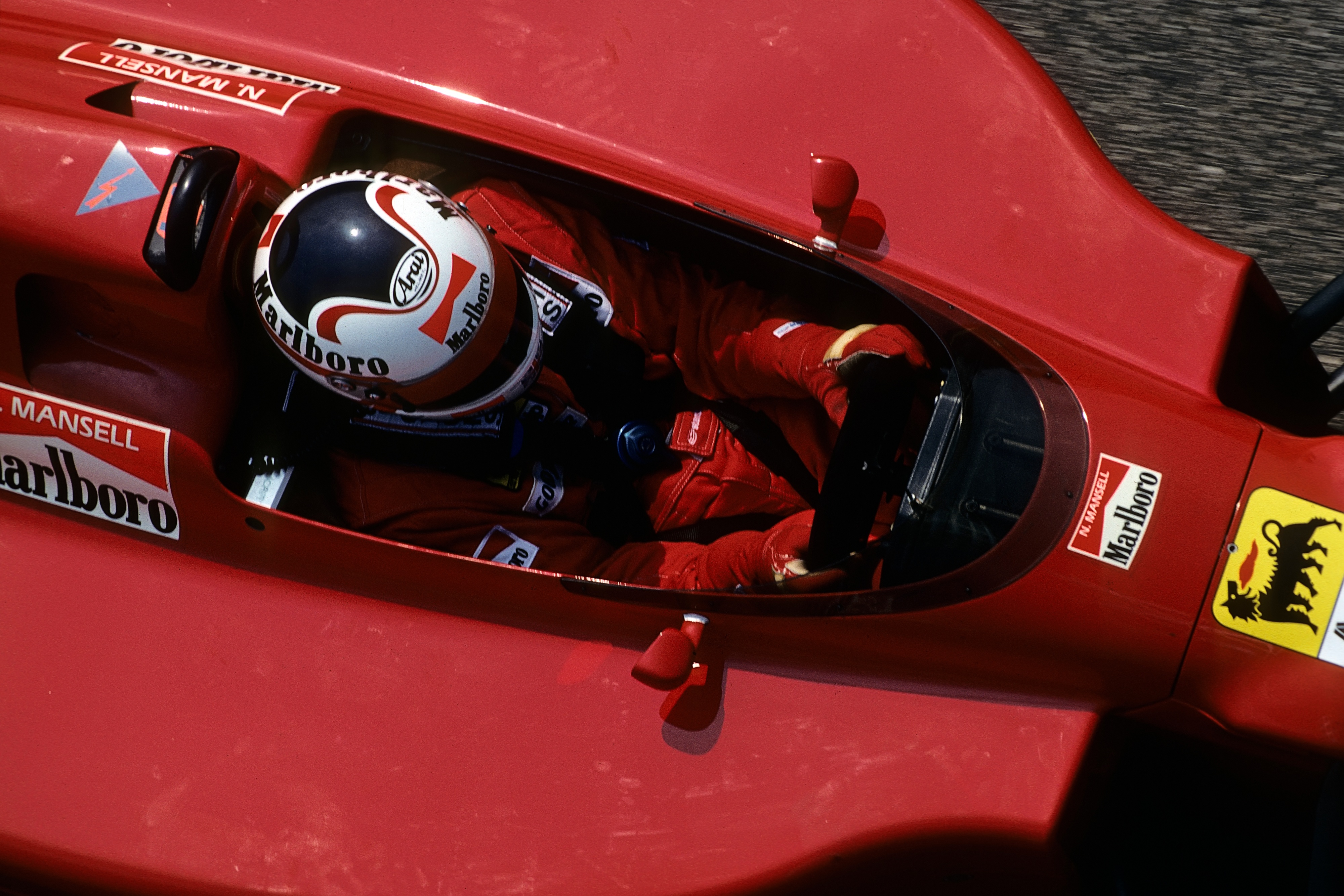 Une voiture de F1 rouge vif prise d'en haut.  La voiture occupe la largeur du cadre de l’image.  À l'intérieur est assis le conducteur, portant un casque blanc et une combinaison de course rouge, les mains sur le volant.  Le casque du conducteur et la voiture sont décorés de logos lisant "Marlboro."